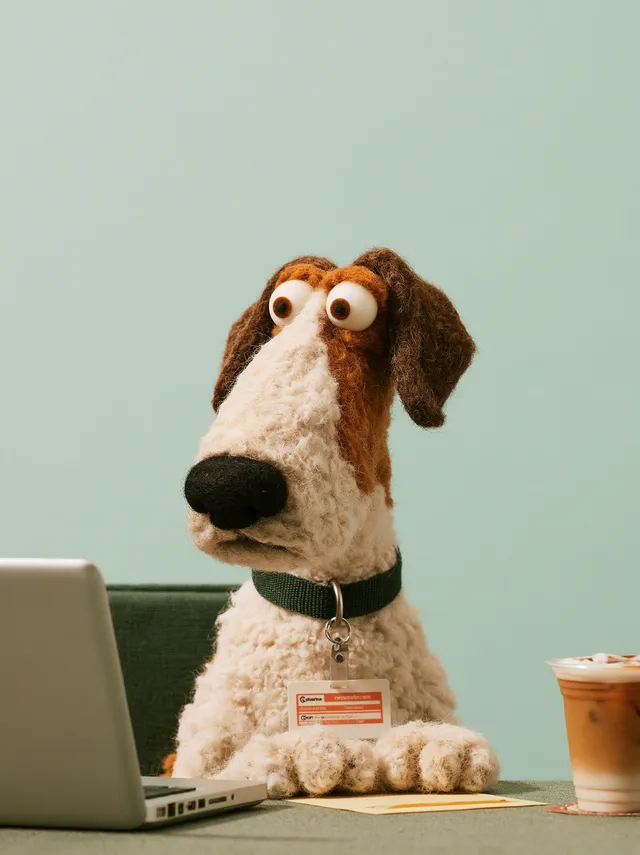 stop-motion felt puppet dog sitting at an office desk explaining something to a cat colleague, wearing a name badge lanyard, laptop and iced