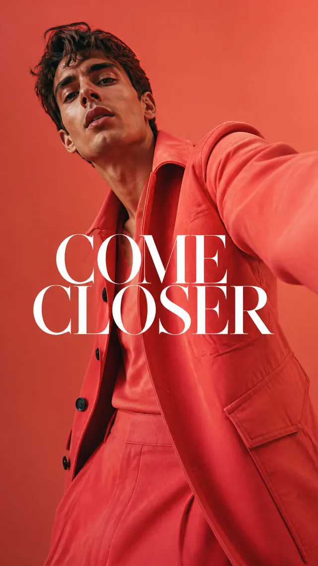 Luxury fashion editorial. Main Subject: Male model wearing a vivid coral jacket. Pose: One shoulder leaning toward the camera crea